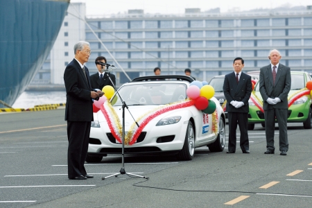 Az óriás komphajó mellett a Mazda elnöke szónokol, aki a megszokottól eltérően gyakran jelenik meg a gyár dolgozói előtt, ezzel is kifejezve a megbecsülésüket.